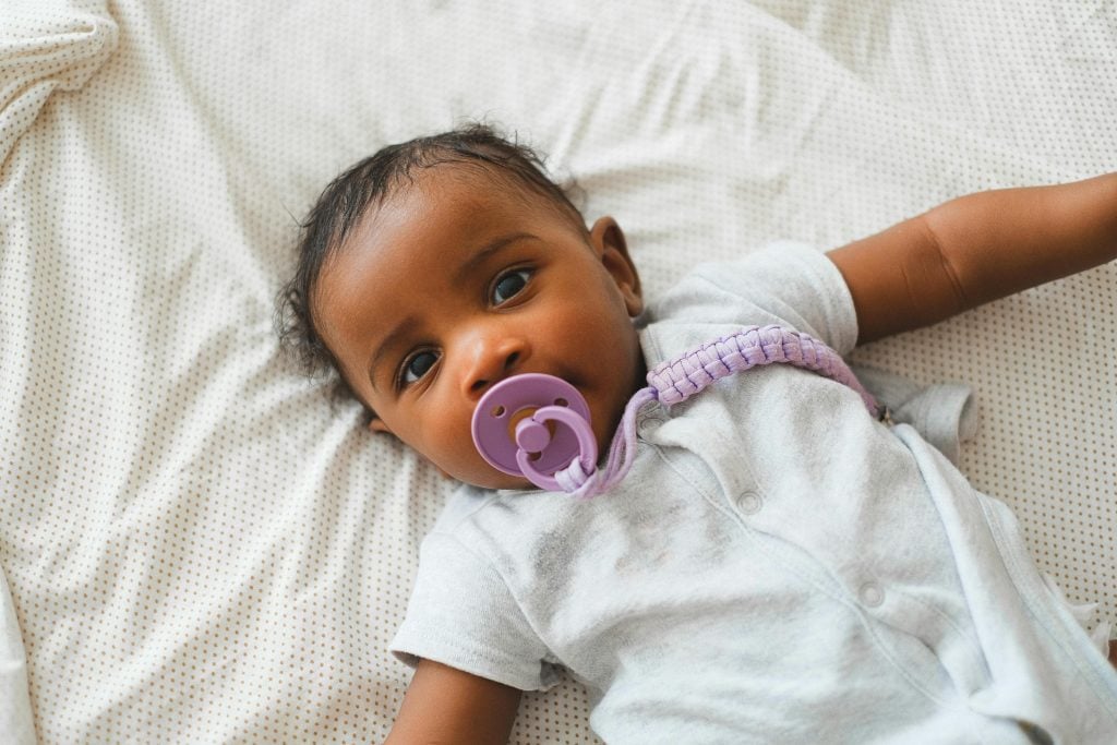 Close-up of a baby in a onesie with a pacifier, lying on a bed.