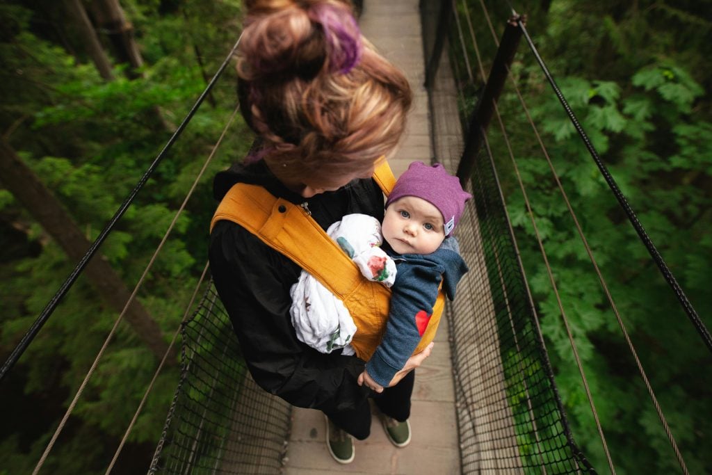 A woman carries her baby over a forest suspension bridge, highlighting maternal care.