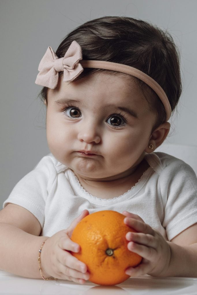 Cute baby with headband holding an orange, showcasing innocent and curious expression.