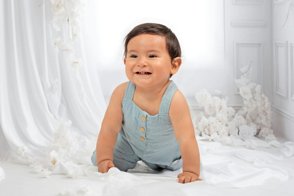 Cute baby in a blue outfit crawling and smiling indoors surrounded by white decor.