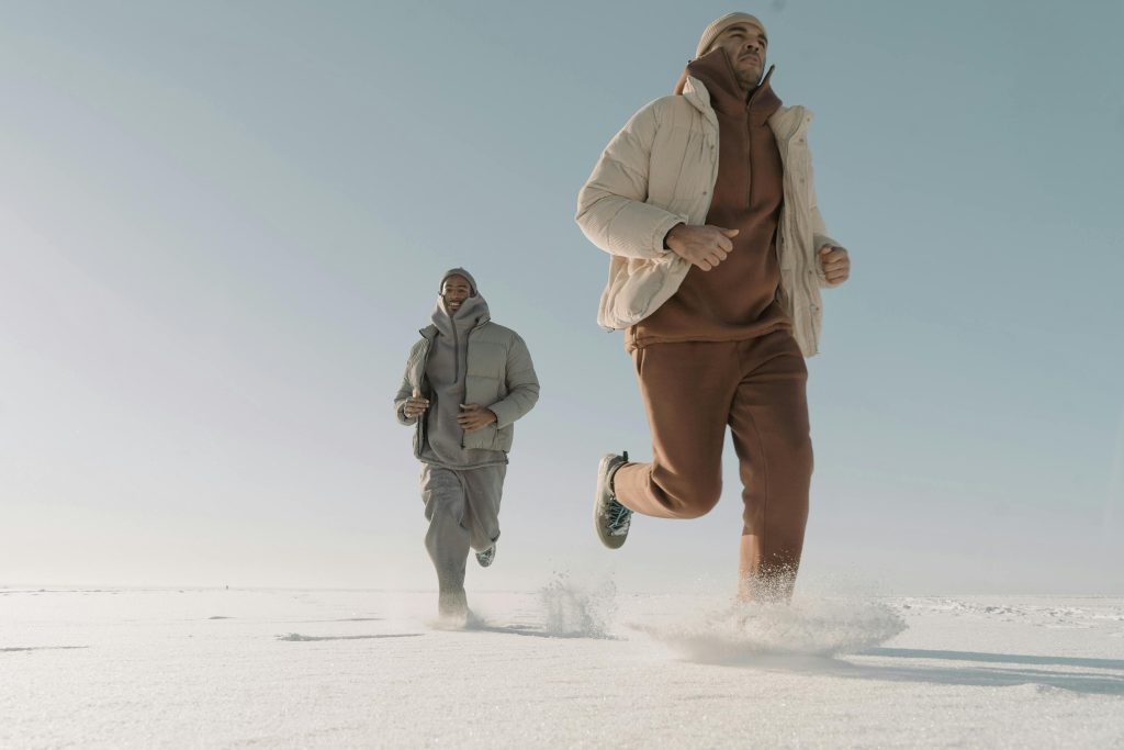 Two men running on a snow-covered ground wearing winter outfits, showcasing fashion and fitness.